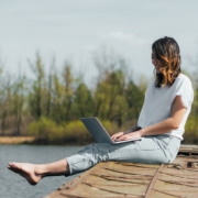 Eine Frau sitzt auf einem Steg am Wasser und schreibt auf einem Laptop, erfolgreich bloggen, entspannt und zielgerichtet bloggen, Spaß am Schreiben