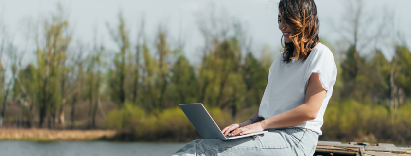 Eine Frau sitzt auf einem Steg am Wasser und schreibt auf einem Laptop, erfolgreich bloggen, entspannt und zielgerichtet bloggen, Spaß am Schreiben